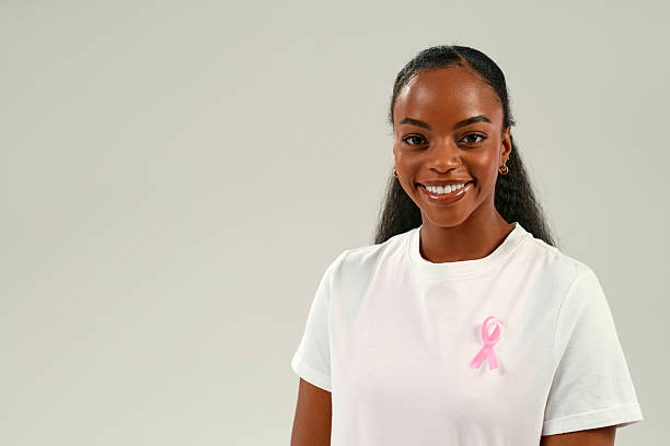 istockphoto-2223578229-612x612 Portrait of smiling African American woman in white t-shirt with pink ribbon pinned on chest, symbolizing breast cancer awareness, hope, and healthcare support concept.
